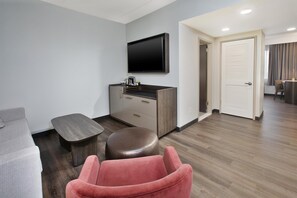 A modern hotel room with a flat-screen TV mounted on the wall, a small wooden desk with a coffee maker, a cushioned bench, and a red chair.