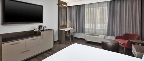 A hotel room with a large bed, a desk with a chair, a flat-screen TV, a coffee maker, and a window with curtains.