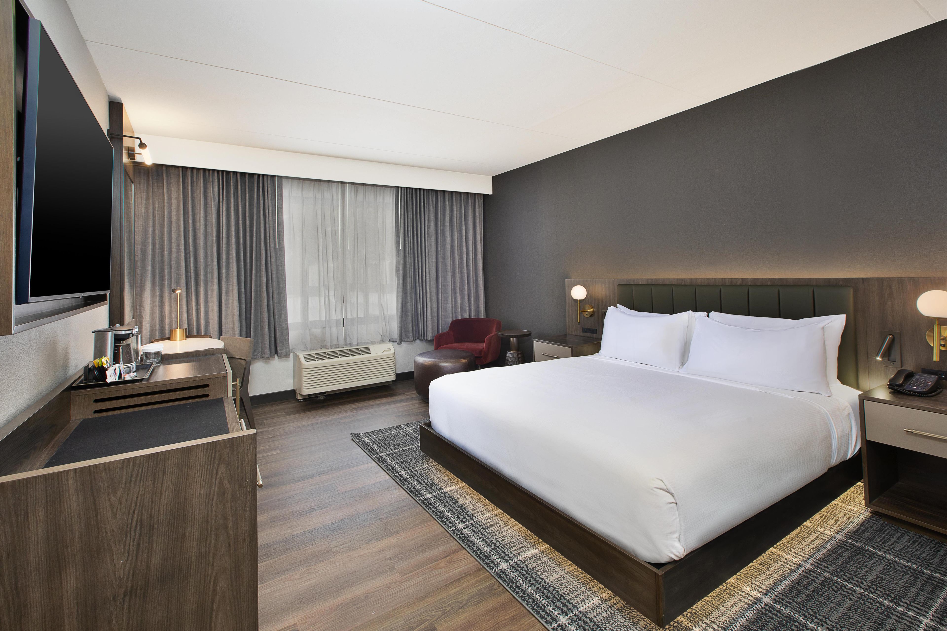 A hotel room with a large bed, a desk with a flat-screen TV, a red chair, a bedside table with a lamp, and a large window with curtains.