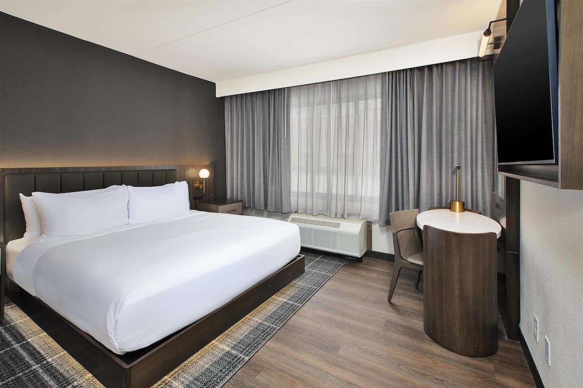 Ein Hotelzimmer mit einem großen Bett, einem Schreibtisch, einem Sessel, einem Fernseher und einem Fenster mit Vorhängen.