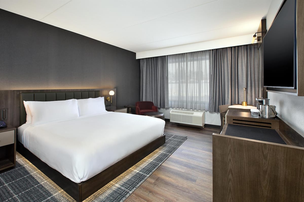 Ein modernes Hotelzimmer mit einem großen Bett, einem Schreibtisch, einem Flachbildfernseher, einem Sessel und einem Fenster mit Vorhängen.