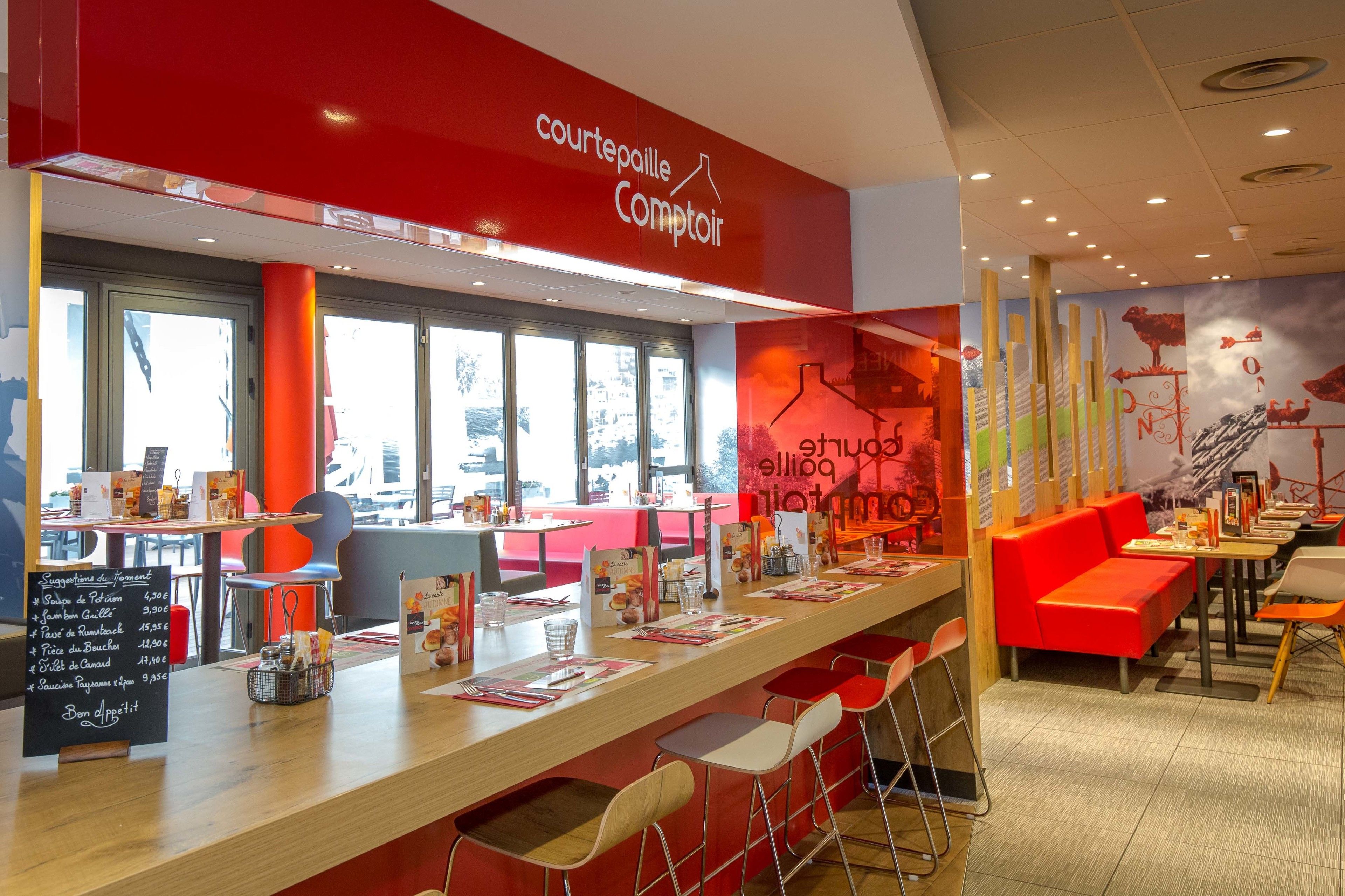 A restaurant with a red and white color scheme, featuring a long counter with stools, a menu board, and a mural on the wall.