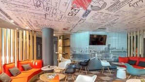 A modern lounge with orange sofas, white and blue chairs, a cityscape mural, and a large TV screen. - ibis Marseille Timone (Marseille)