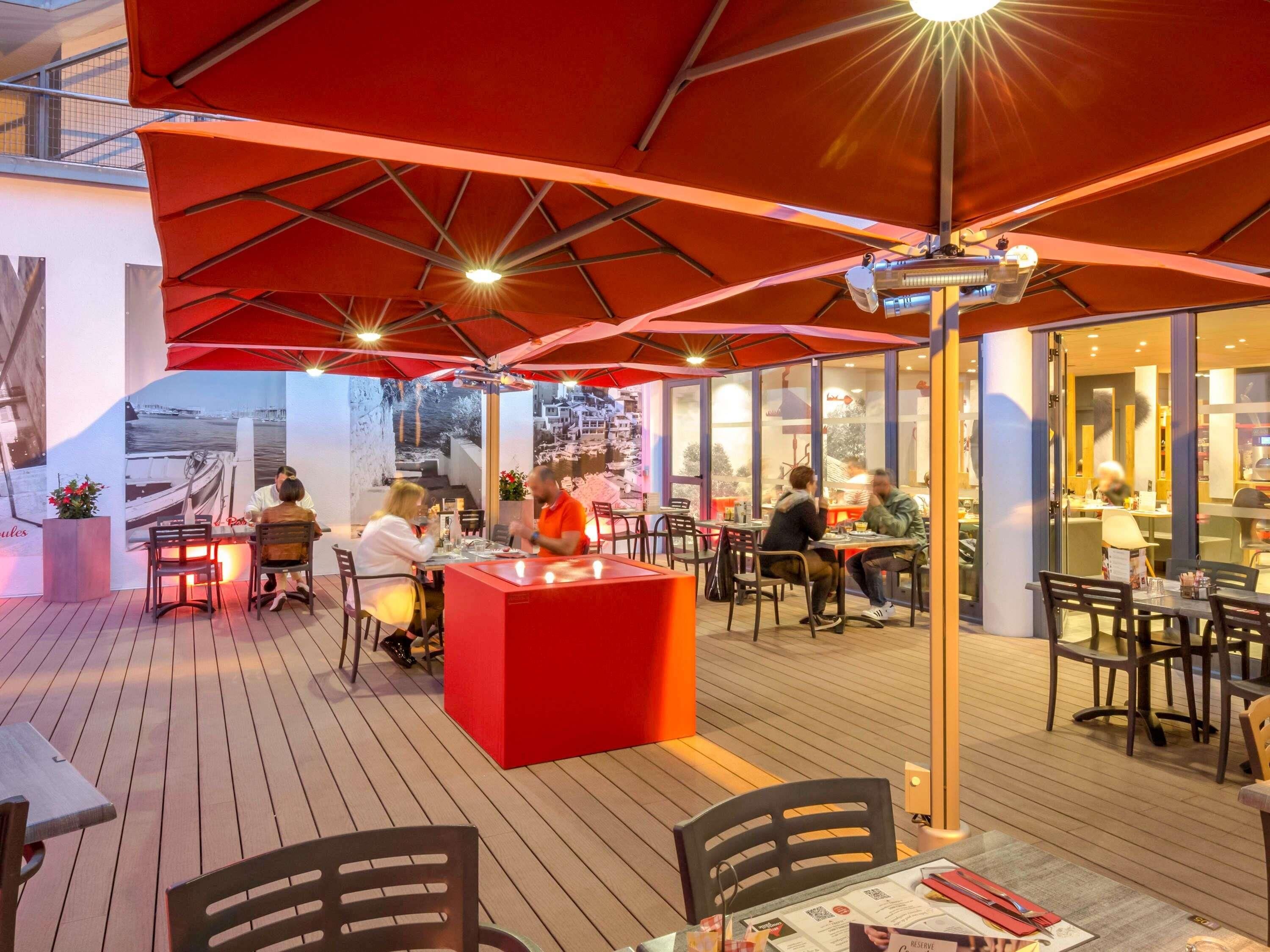 An outdoor dining area with tables and chairs, a red counter, and a view of the cityscape through large windows.
