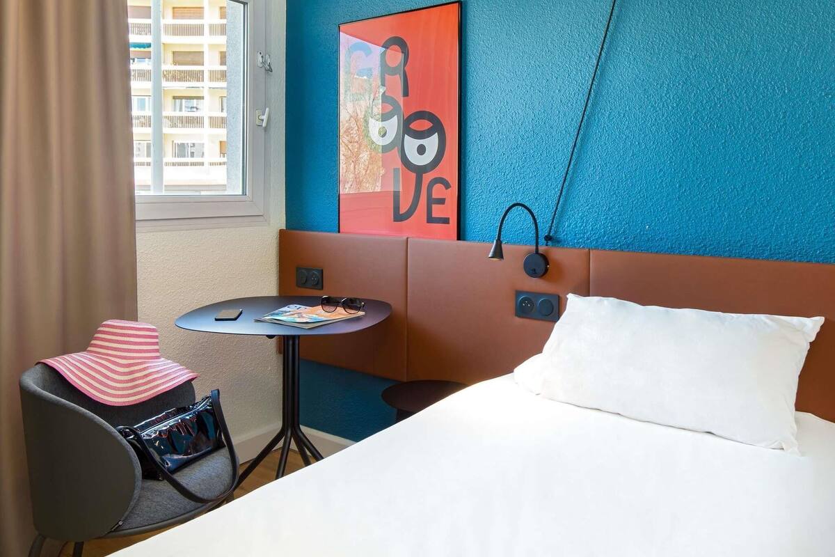 A hotel room with a bed, a small table with a magazine, a chair, a handbag, a window with curtains, and a wall with a colorful artwork.