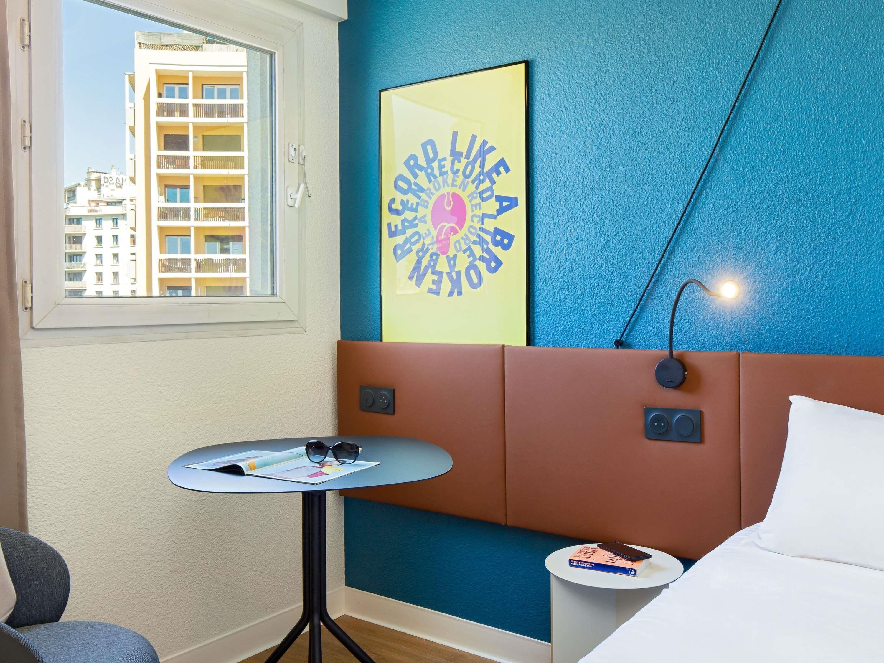 Habitación de hotel con cama, una mesita con una revista y gafas de sol, una lámpara de pared y un cartel.
