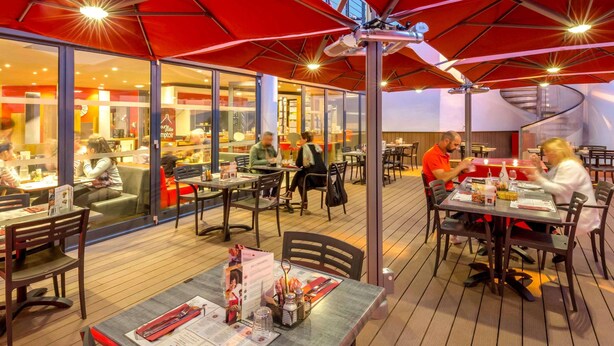 An outdoor dining area with tables and chairs, covered by red umbrellas, and a glass wall separating the dining area from the restaurant inside. - ibis Marseille Timone (Marseille)