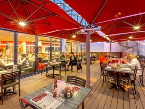 An outdoor dining area with tables and chairs, covered by red umbrellas, and a glass wall separating the dining area from the restaurant inside. - ibis Marseille Timone (Marseille)