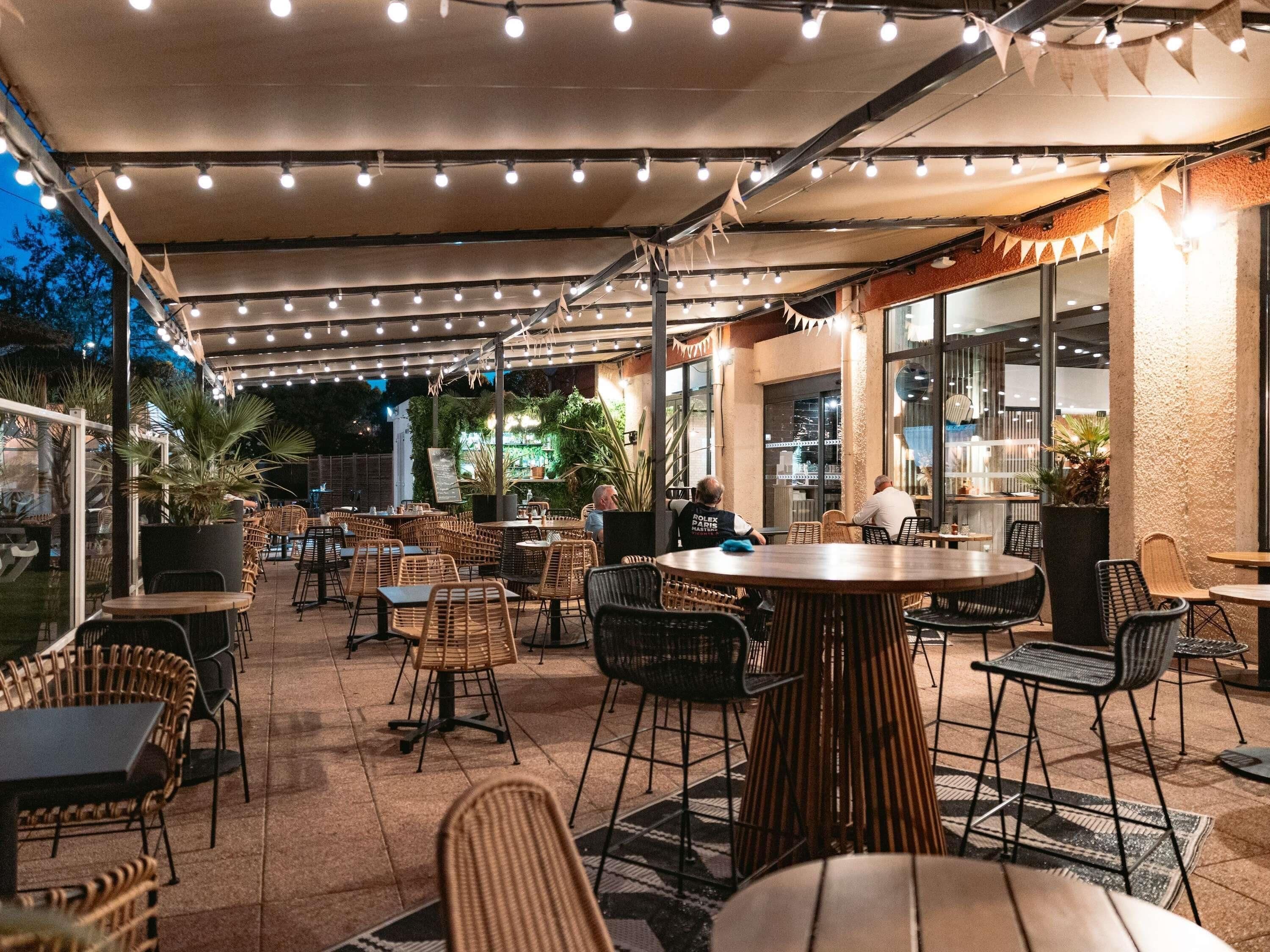 Un café al aire libre con mobiliario de mimbre, luces colgantes y vistas de la ciudad.