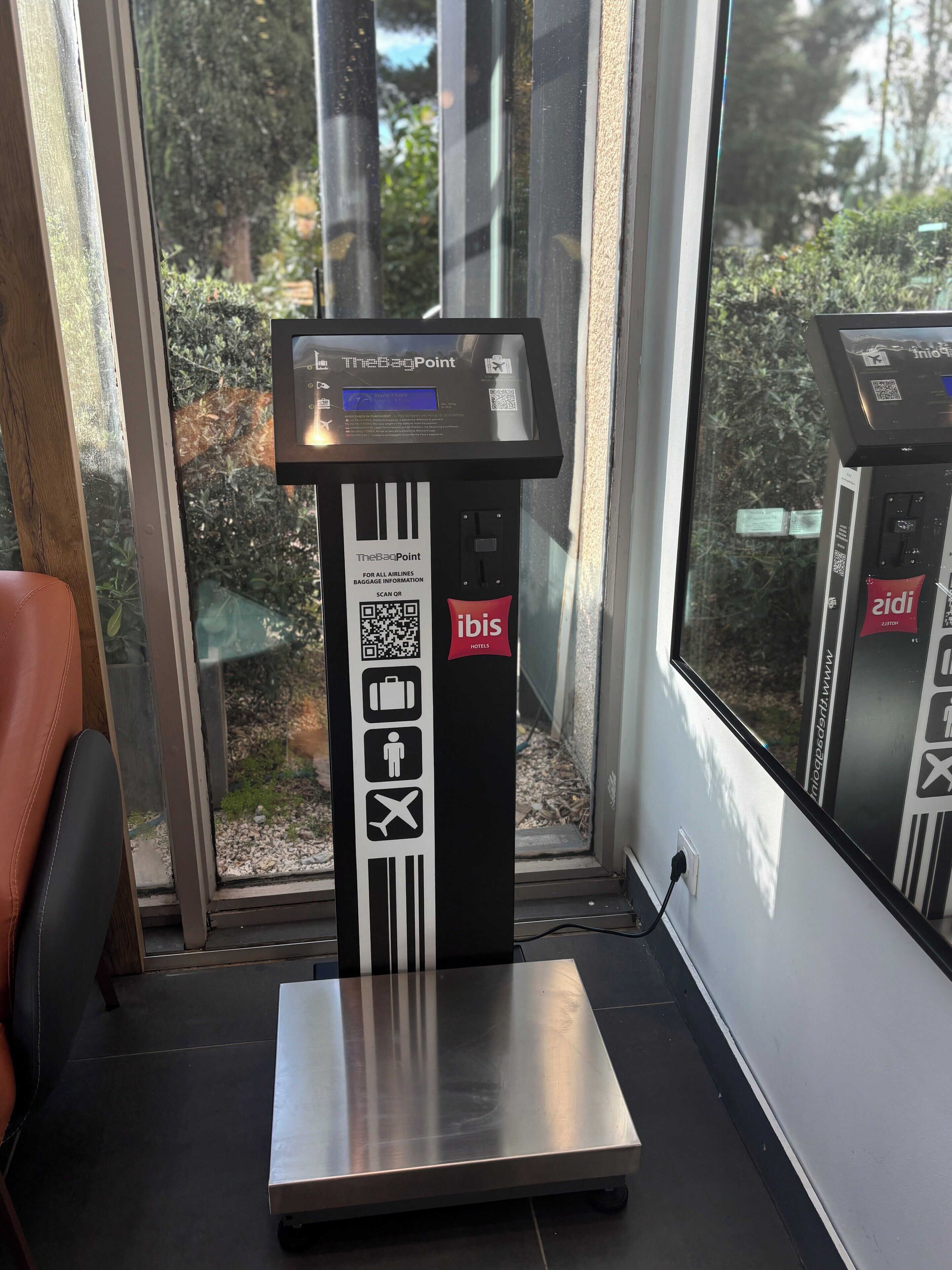 Balanza de equipaje con pantalla digital, un código QR e iconos de maletas, una persona y un avión, que indica que sirve para consultar información de equipaje.