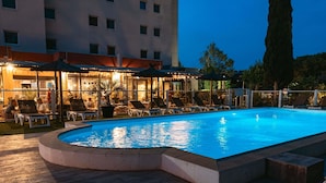 Una piscina con sillas de descanso y sombrillas, rodeada por un edificio de varios pisos y ventanas.