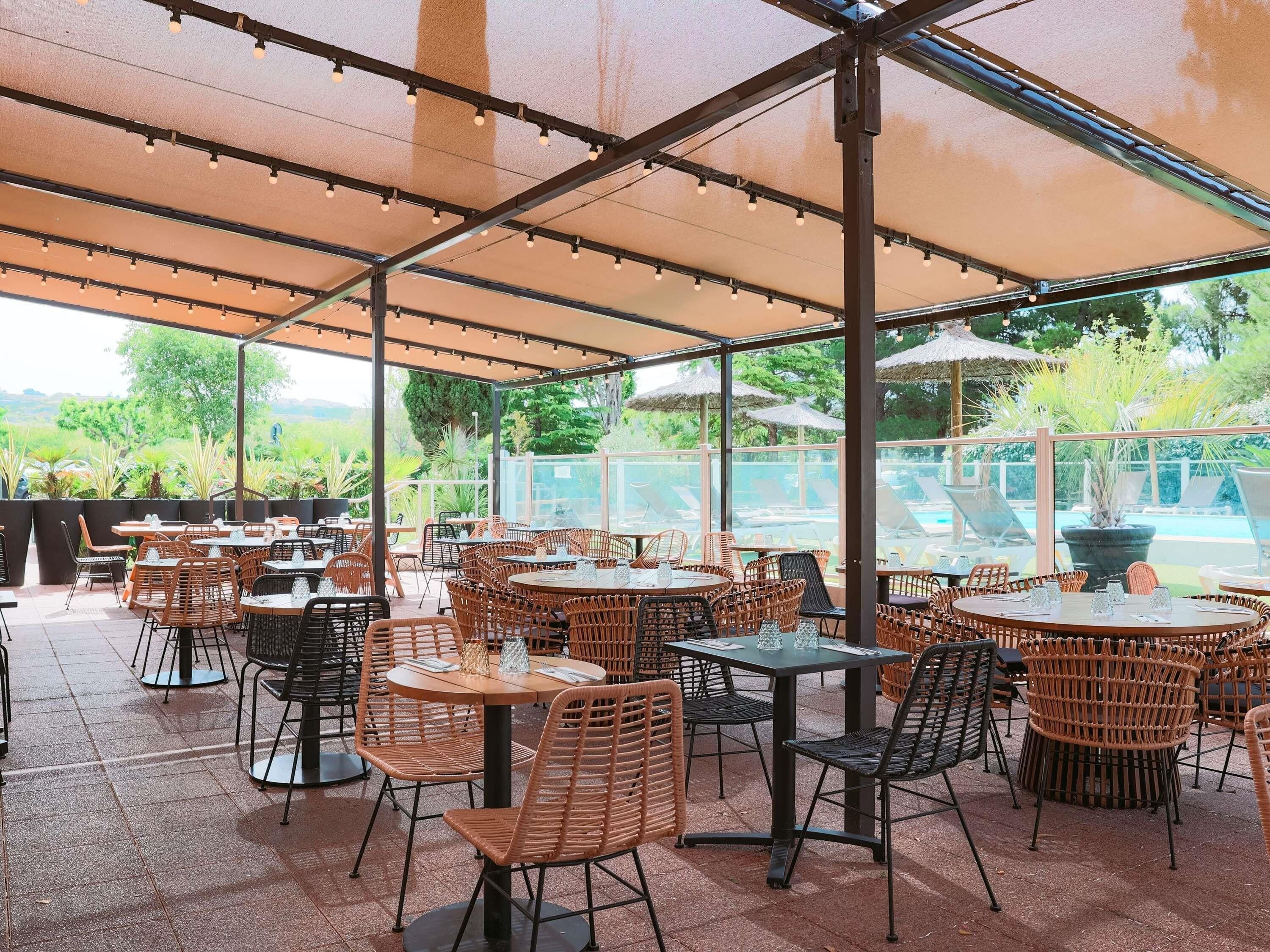 An outdoor patio with round tables and wicker chairs, a pool in the background, and a covered area with string lights.