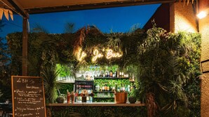 Un bar al aire libre con un letrero de neón, rodeado de abundante follaje, y un menú a la vista.