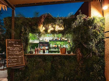 An outdoor bar with a neon sign, surrounded by lush greenery, and a menu board visible.