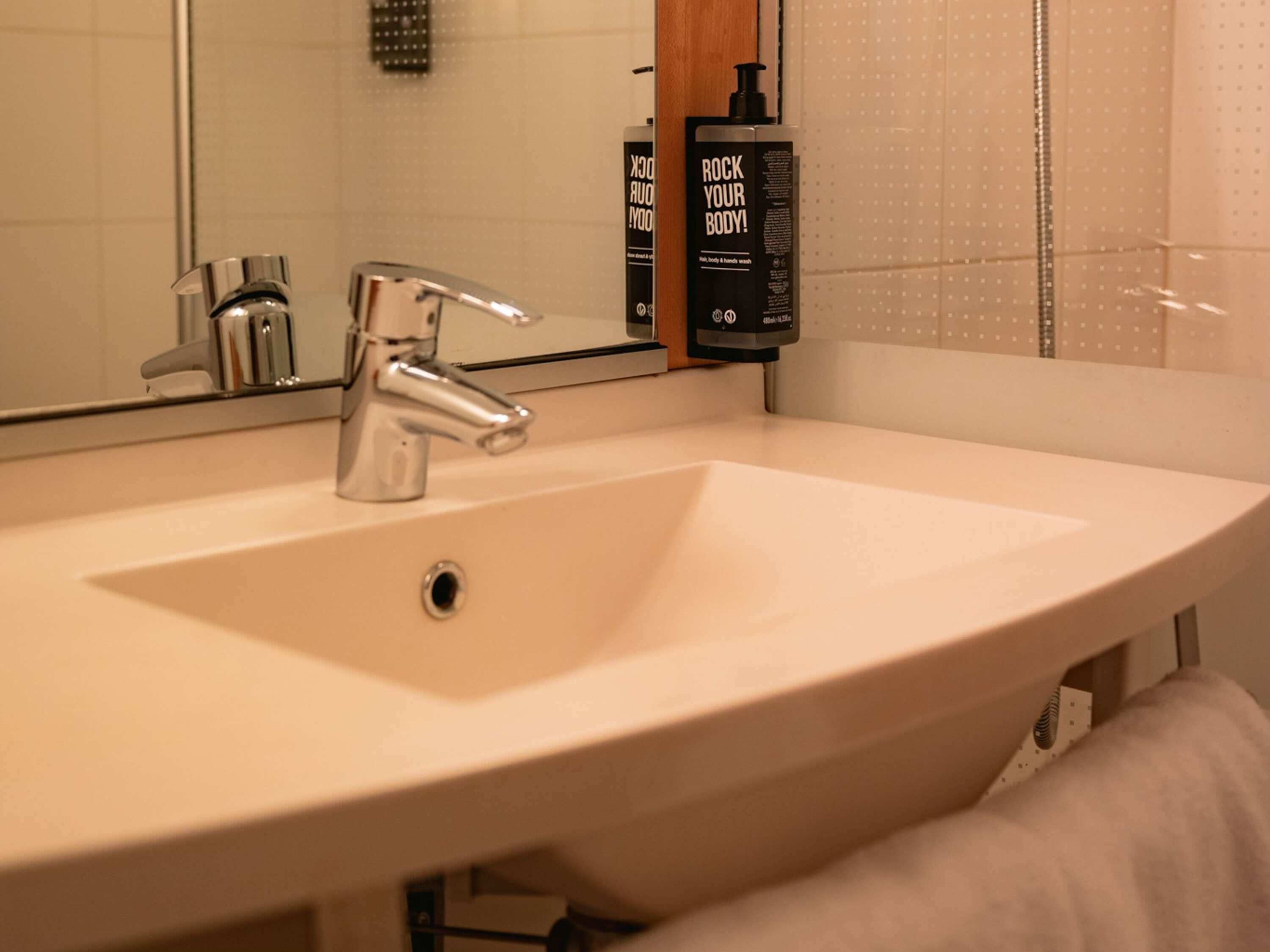Une salle de bain avec un lavabo blanc, un robinet chromé et un miroir. Deux flacons de gel douche sont posés sur le mur.
