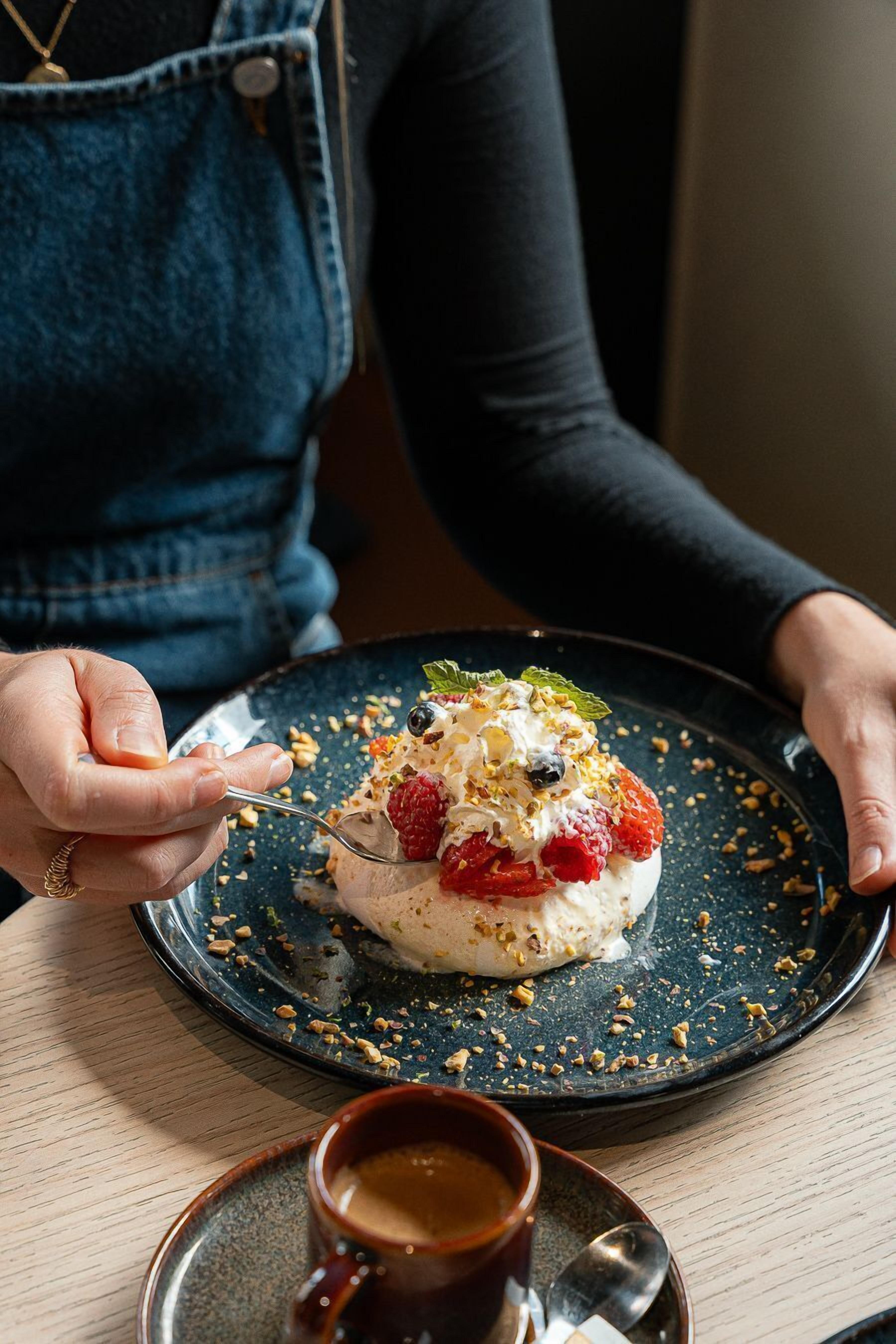 A person holding a plate with a dessert topped with berries and nuts, and a cup of coffee on the table.