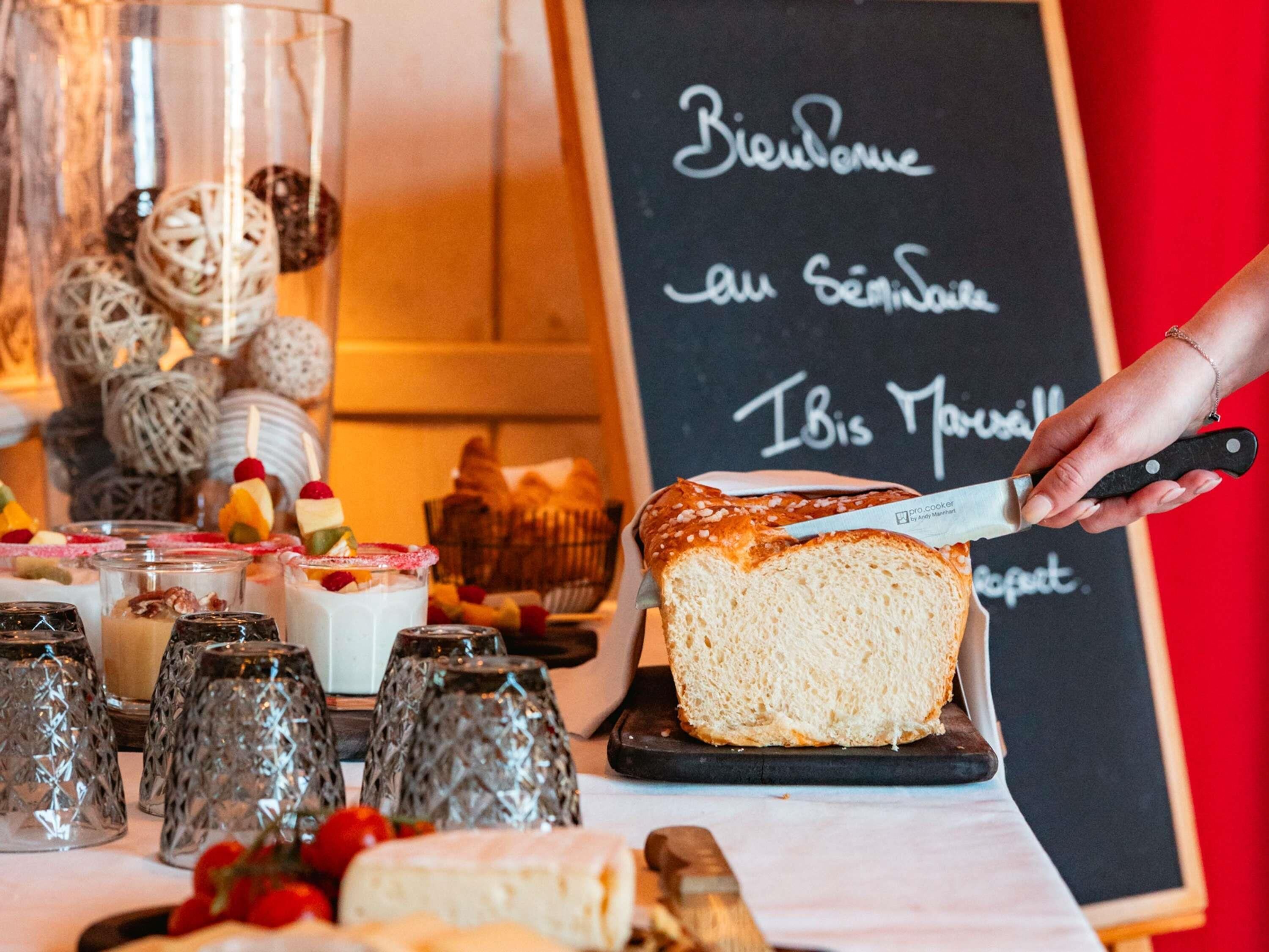 Un comptoir de boulangerie où l’on tranche un pain, avec une variété de pâtisseries et un tableau noir sur lequel sont écrits des messages de bienvenue.