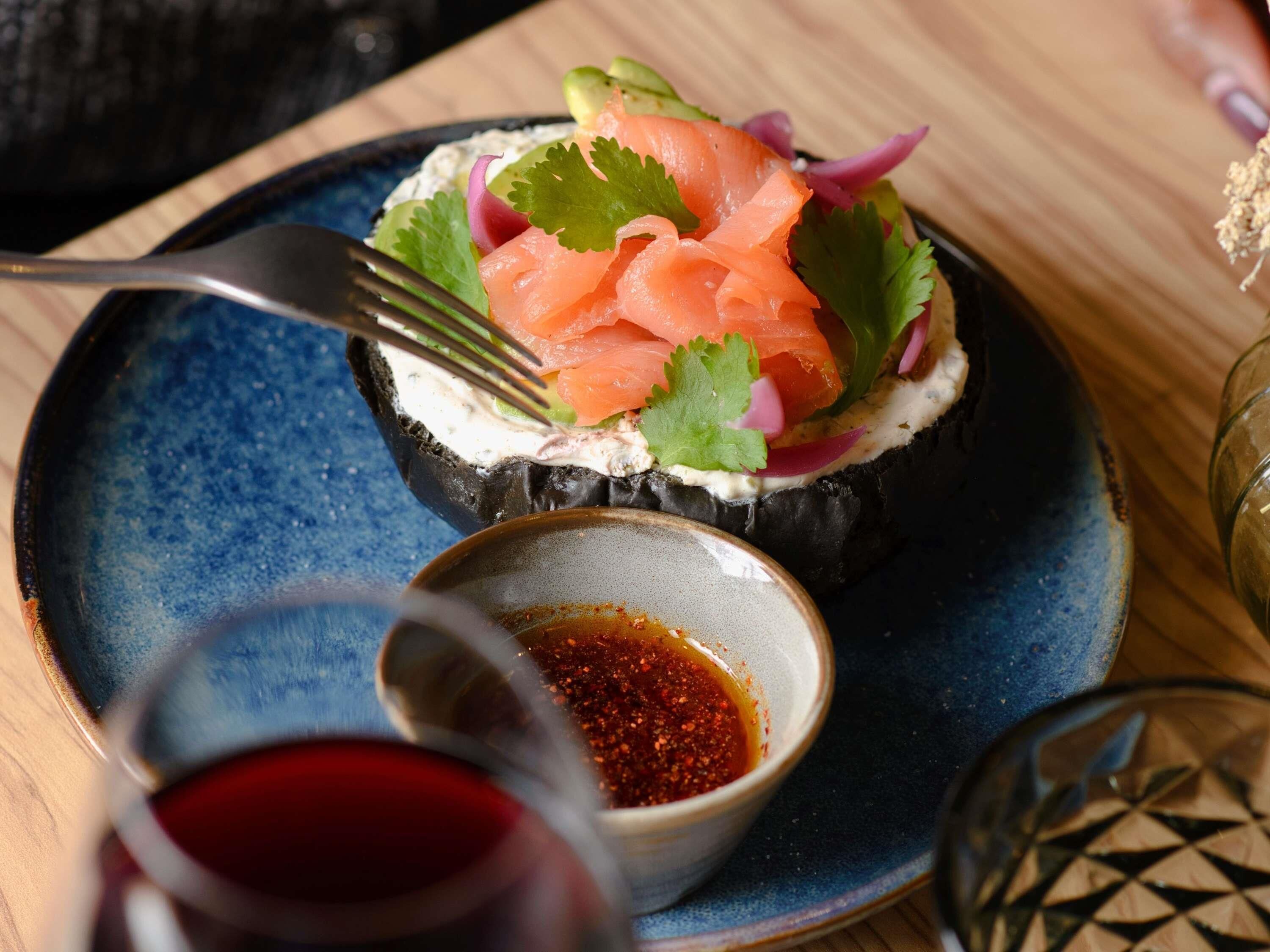 Un plato con salmón, crema y verduras sobre un plato negro, acompañado de un recipiente pequeño con salsa roja y un vaso de líquido rojo.