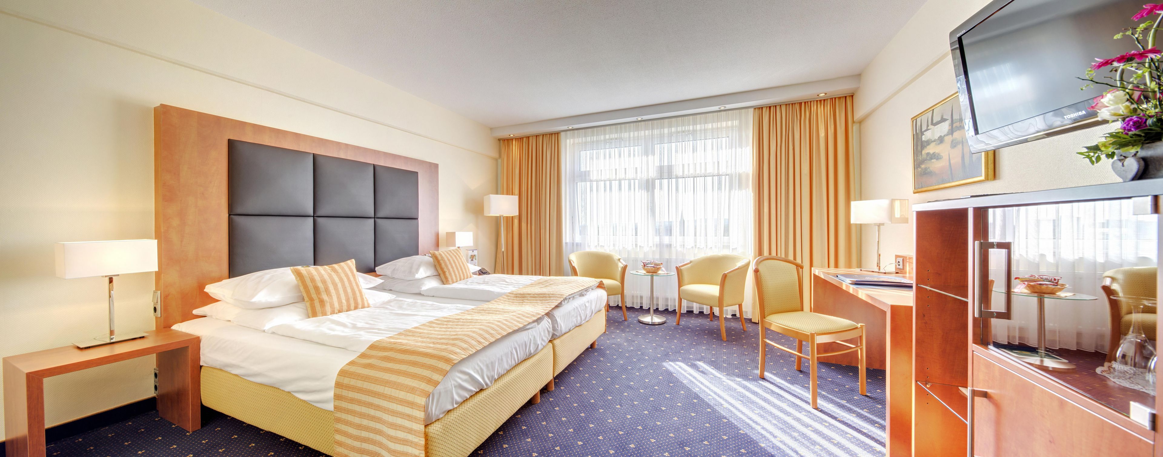 A hotel room with a double bed, two bedside tables, a desk with a chair, a TV, and a window with curtains.