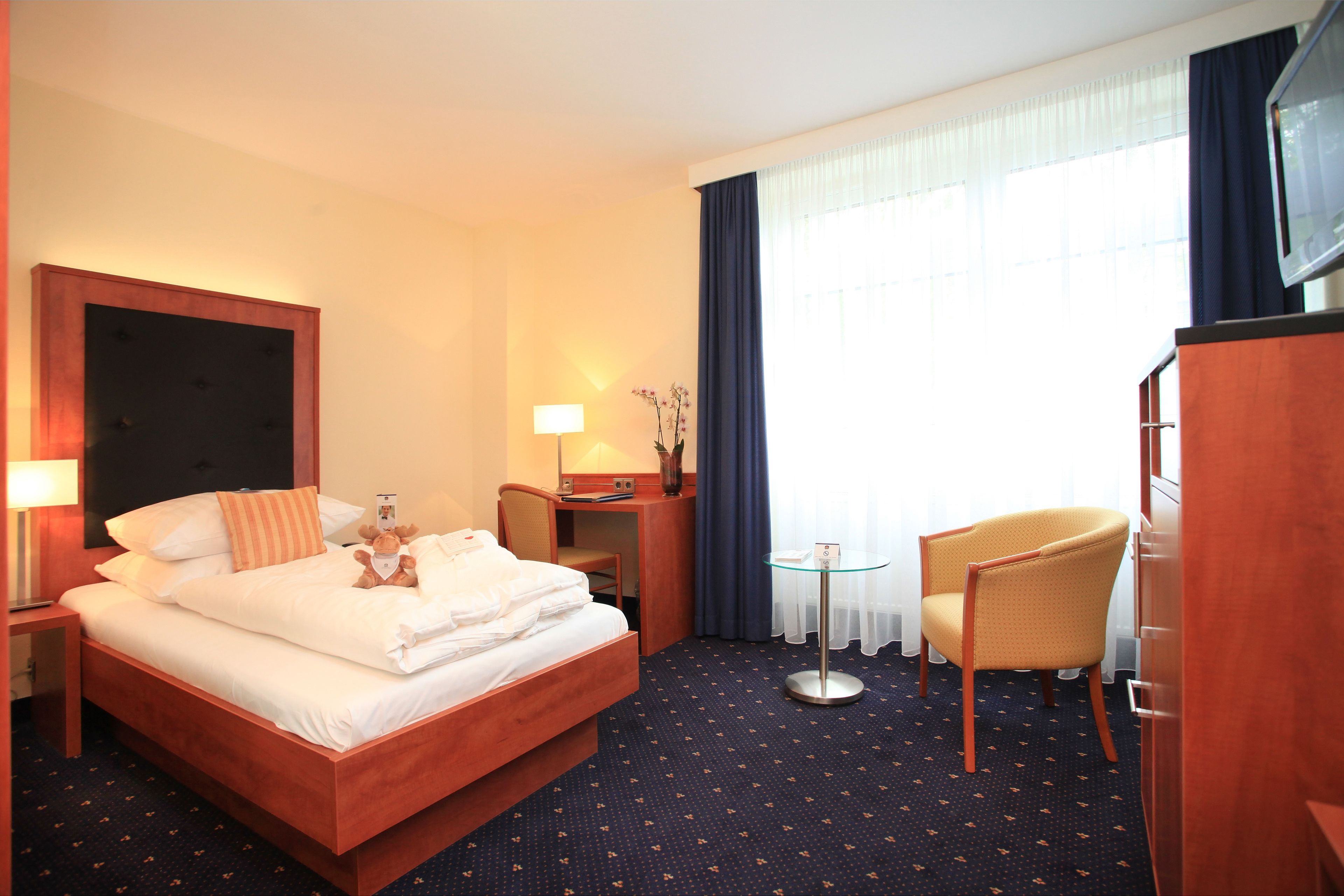A hotel room with a bed, a desk, a chair, a TV, and a small table with a lamp.