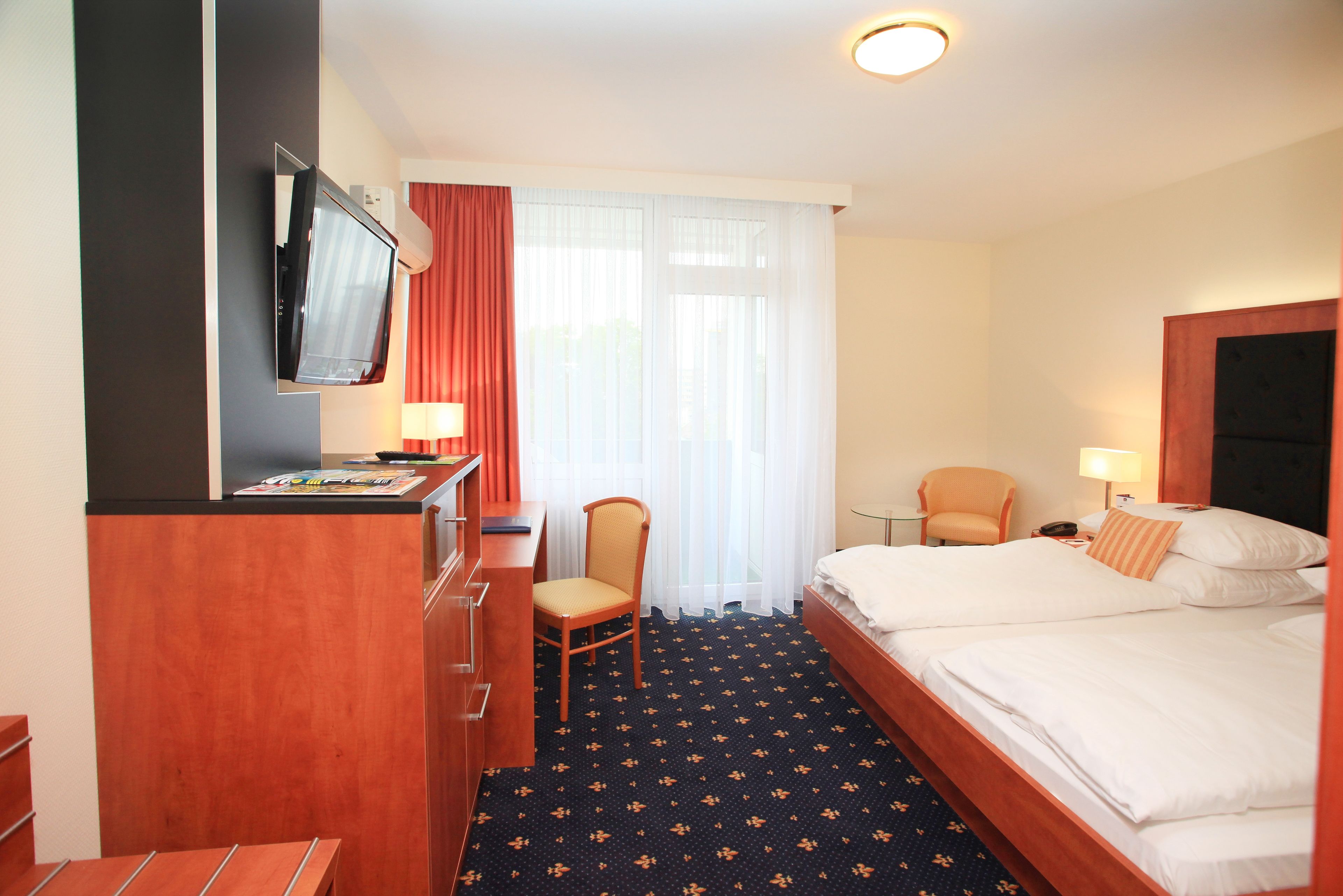 Habitación de hotel con cama, escritorio con silla, televisor montado en la pared y ventana con cortinas.