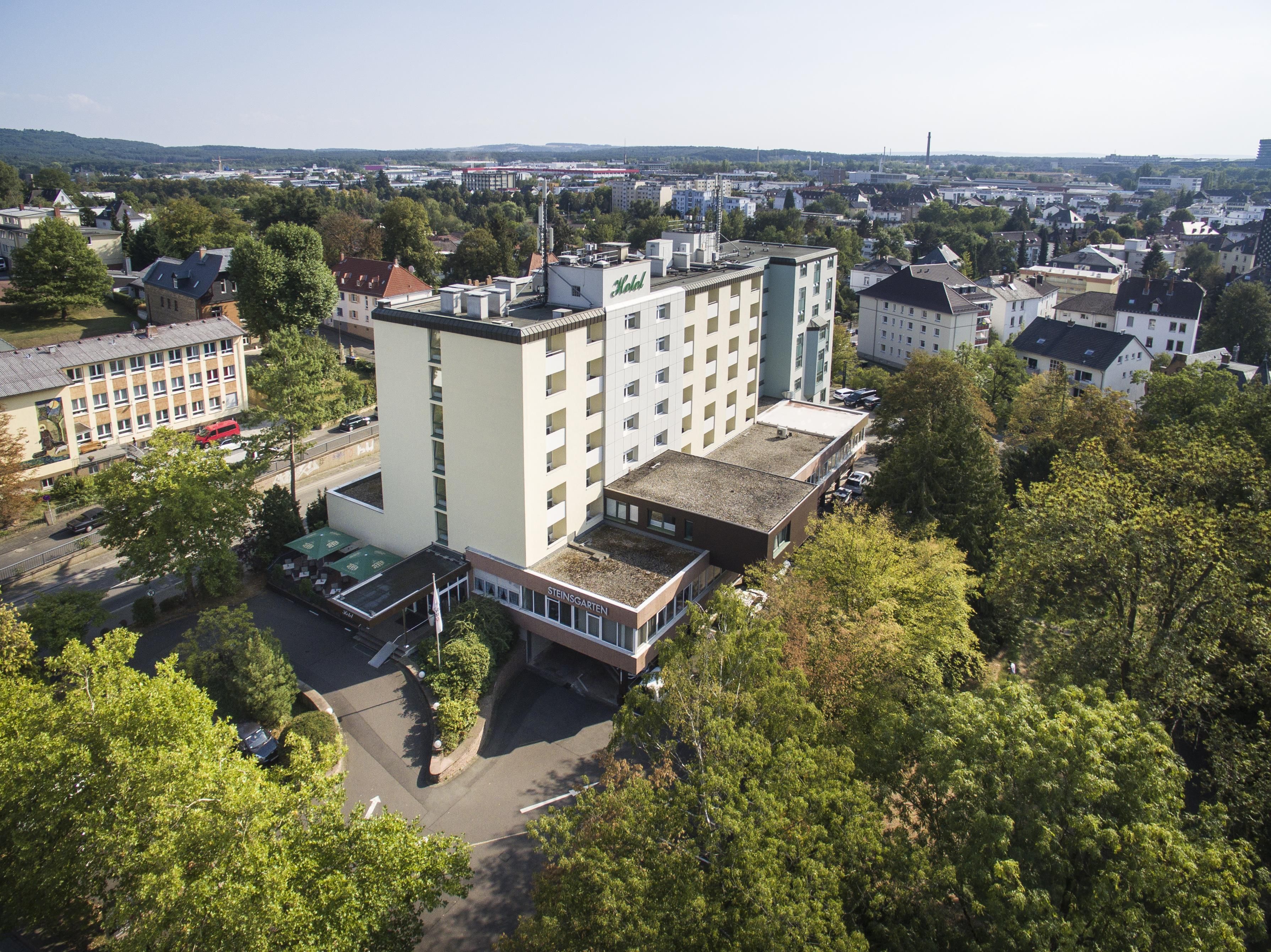 A multi-story building with a flat roof, surrounded by trees and other buildings.