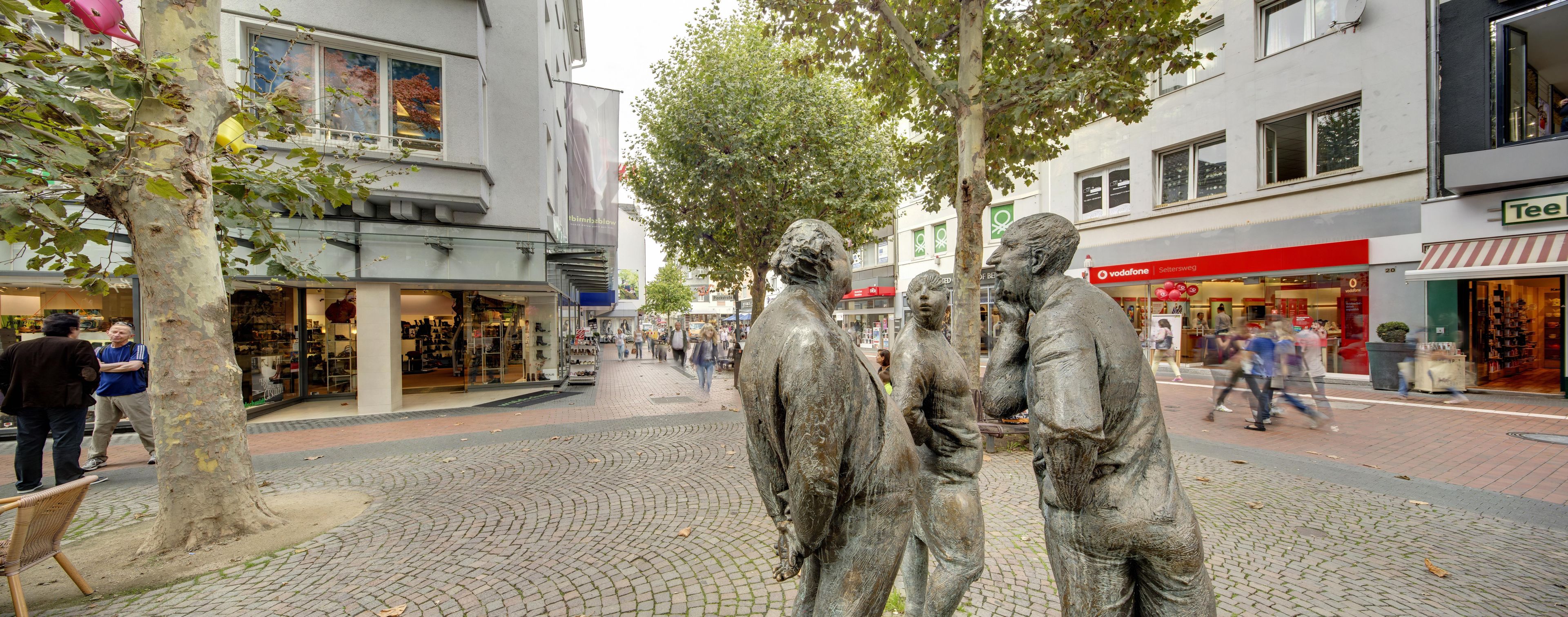 A public square with a statue of three figures, shops, and a Vodafone store.