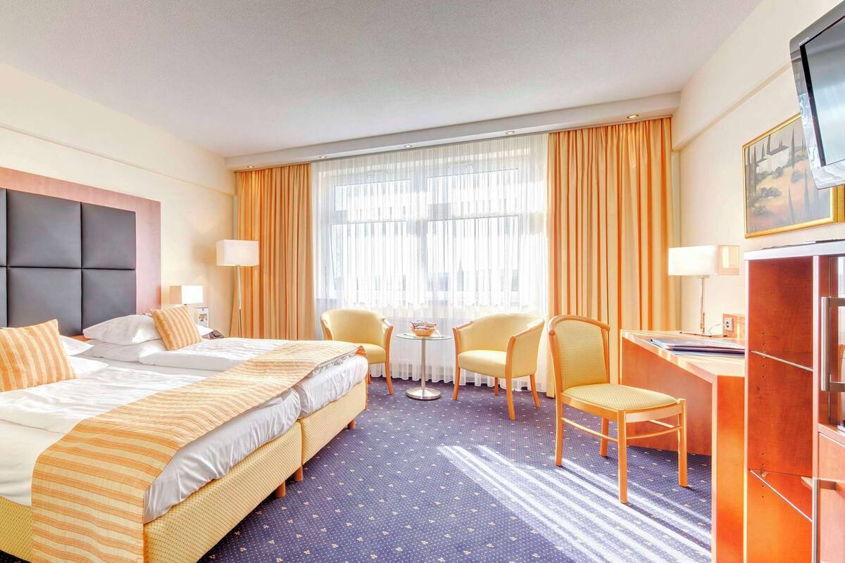 A hotel room with a large bed, two bedside tables, a desk with a chair, a television, and a window with curtains.