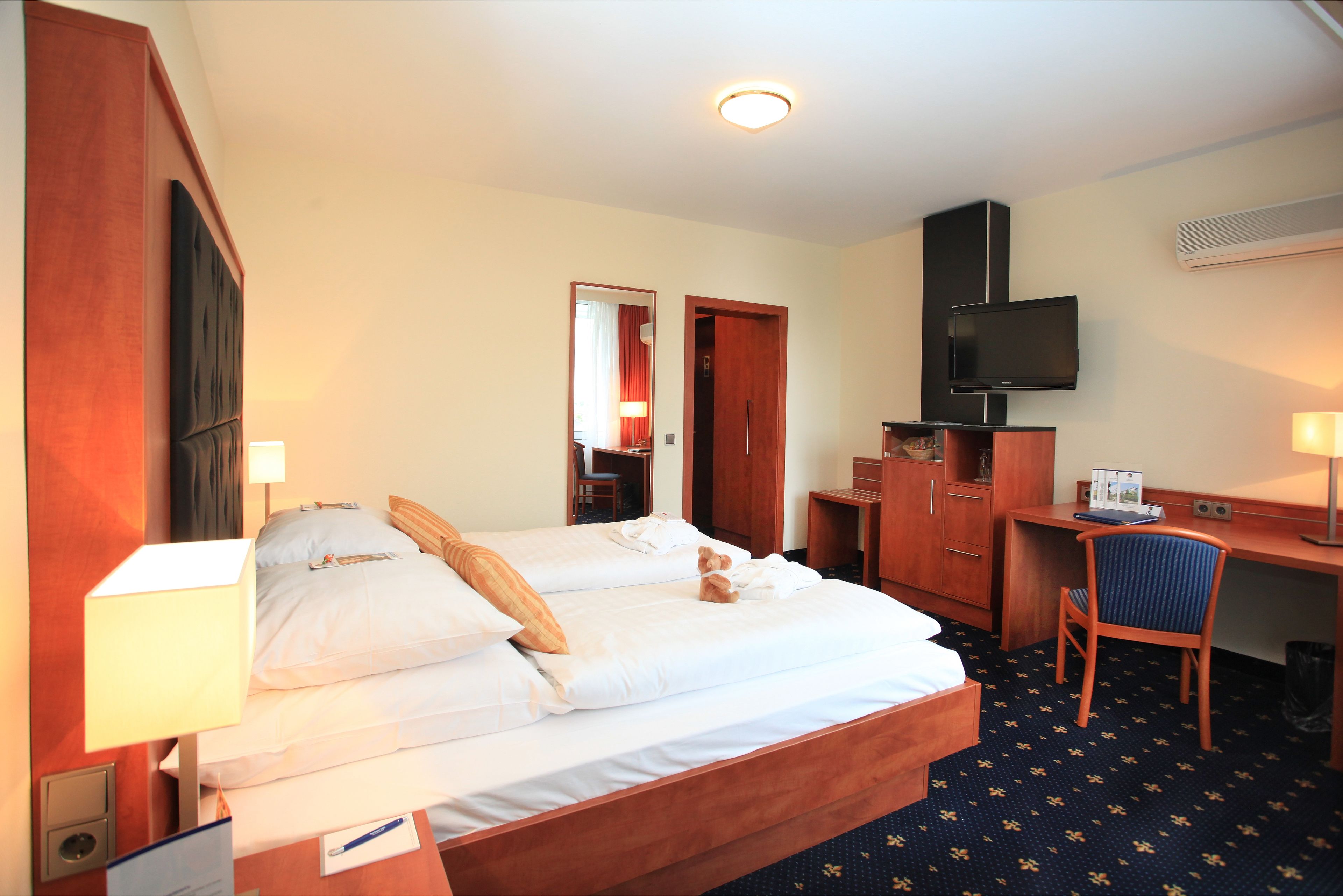 Ein Hotelzimmer mit zwei Betten, einem Schreibtisch, einem Sessel, einem Fernseher und einem Spiegel.