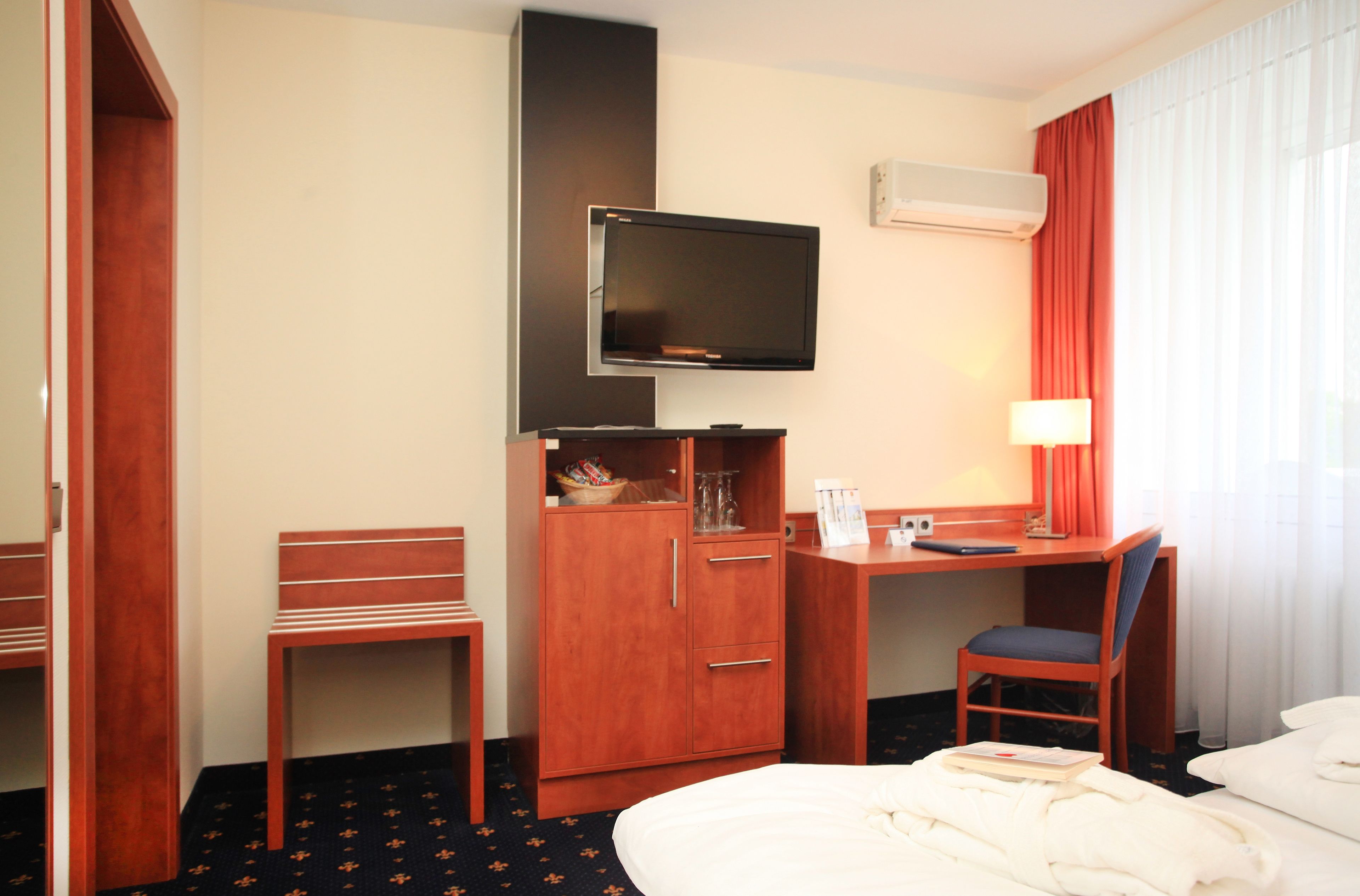 A hotel room with a bed, a desk with a chair, a television, a wardrobe, and a window with curtains.