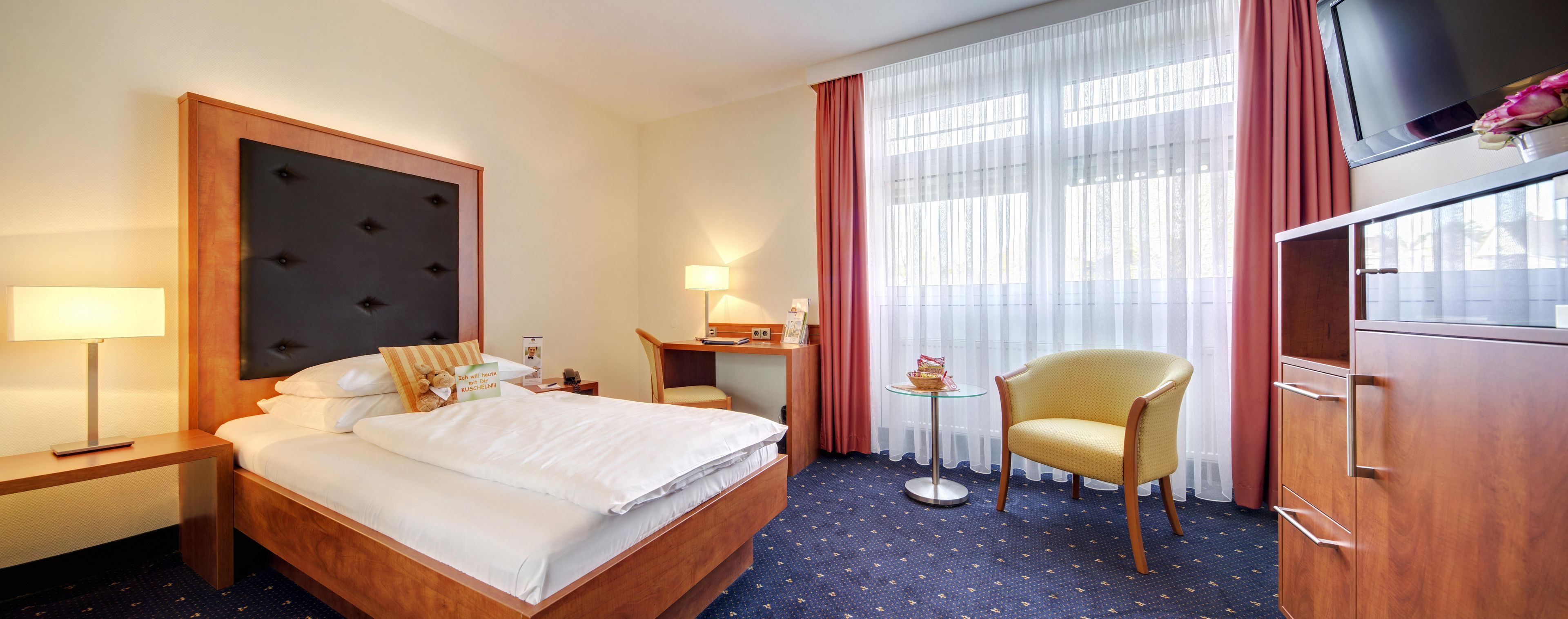 A hotel room with a bed, bedside table, lamp, chair, desk, and a television.