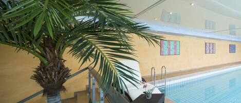 Un área de piscina interior con una palmera, una pequeña mesa con un jarrón y una silla.