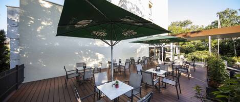Eine Außenpatio mit Tischen und Stühlen, grünen Sonnenschirmen und einem weißen Gebäude im Hintergrund.