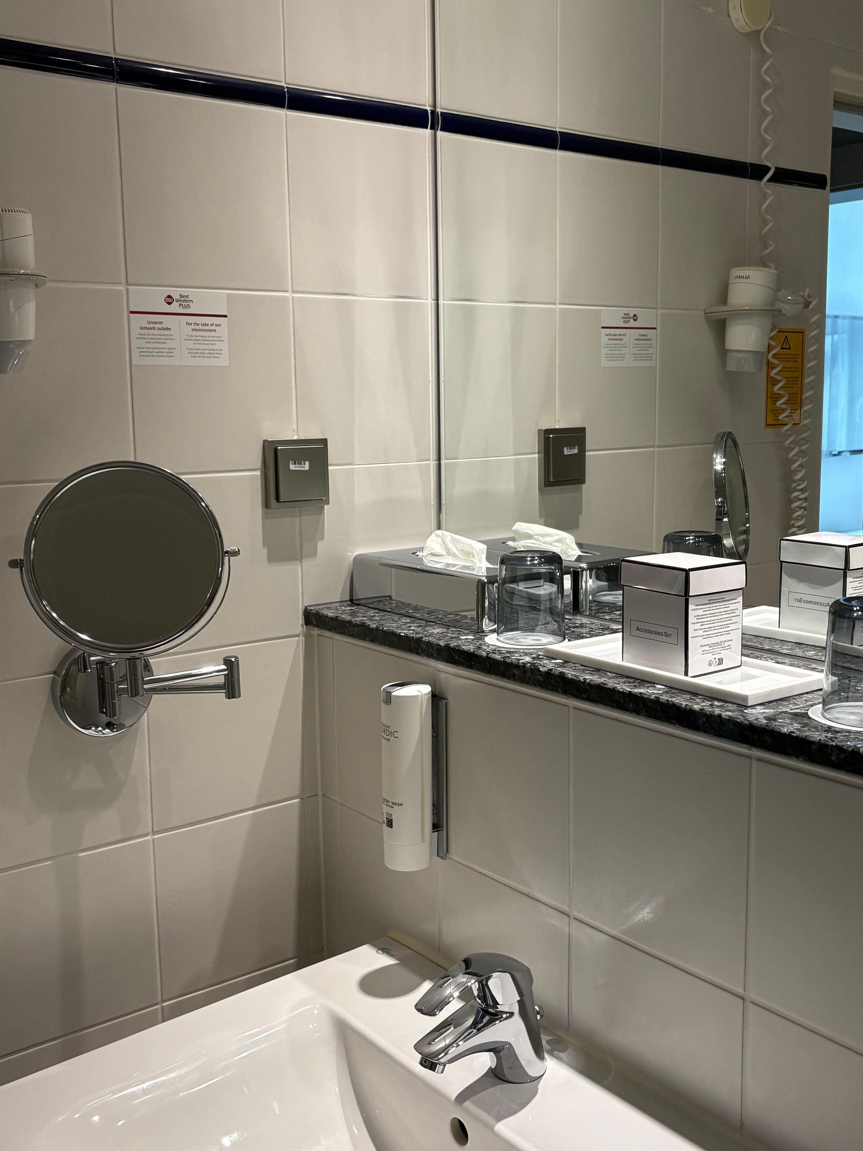 A modern bathroom with a white sink, a mirror, and a toilet. There are toiletries on the counter and a towel dispenser.