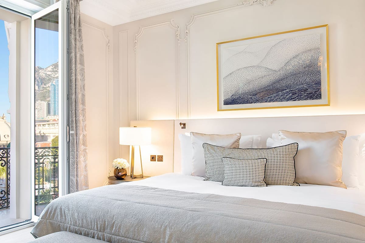 A hotel room with a large bed, a nightstand with a lamp, a framed artwork above the bed, and a balcony with a view of the city.