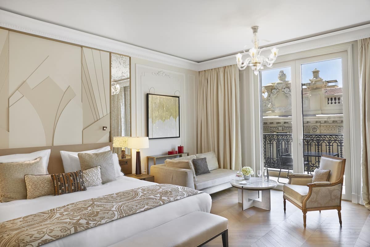 A hotel room with a large bed, a sofa, a chair, a small table, and a balcony with a view of buildings.