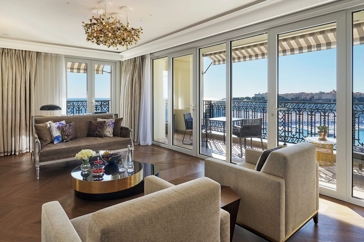 A spacious living room with a sofa, armchairs, and a coffee table. Large windows offer a view of the sea and a balcony with outdoor furniture.