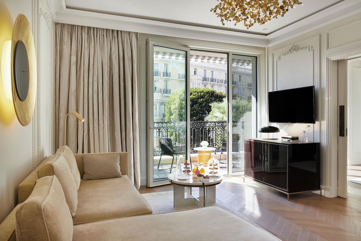 A modern living room with a sofa, a flat-screen TV, a dining table with a glass top, and a balcony with a view of buildings and greenery.