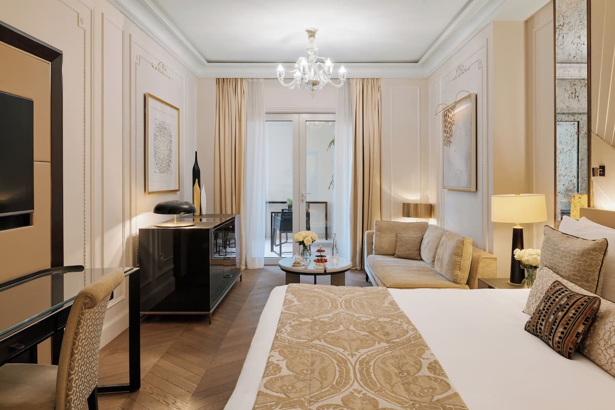 A hotel room with a bed, a desk, a chair, a television, a chandelier, and a view of the outdoors.