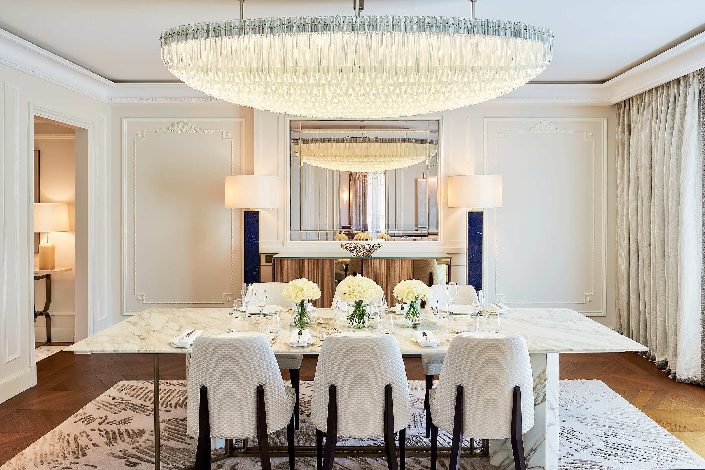 Un comedor formal con una mesa de mármol, sillas blancas con tapicería, una lámpara de araña grande y un espejo sobre la mesa.