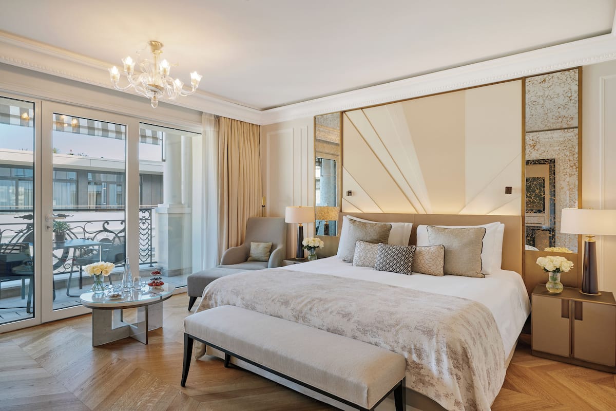 A hotel room with a large bed, a bench, a round coffee table, a chair, a bedside table with lamps, a large mirror, and a view of the outside balcony.