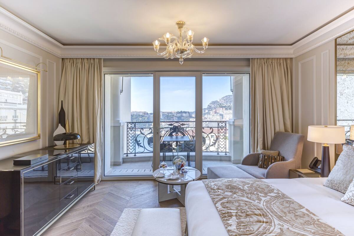 A hotel room with a large bed, a sitting area with a table, a TV, and a balcony with a city view.
