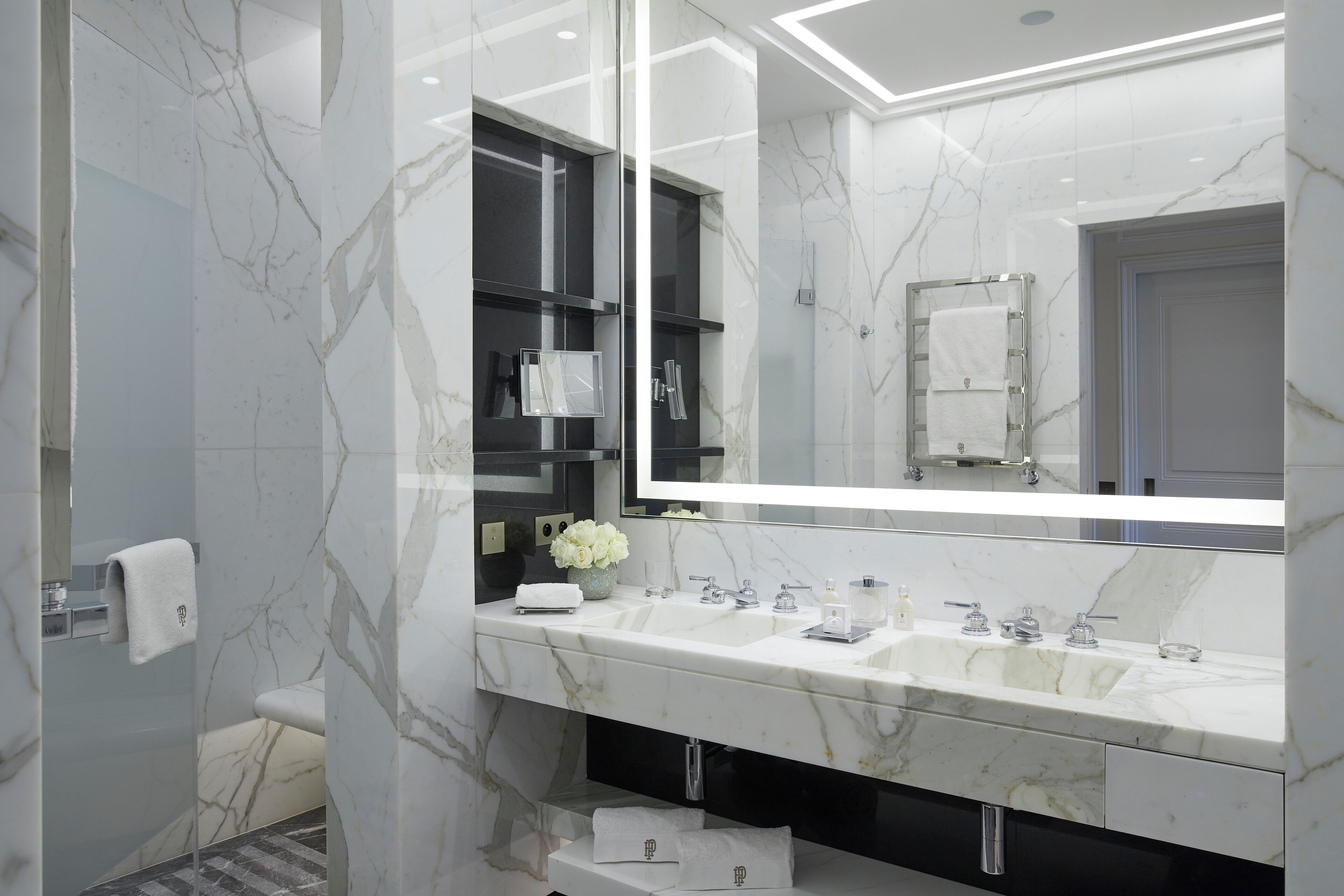 Un baño moderno con paredes de mármol, un espejo grande, lavamanos dobles y mampara de ducha de vidrio.