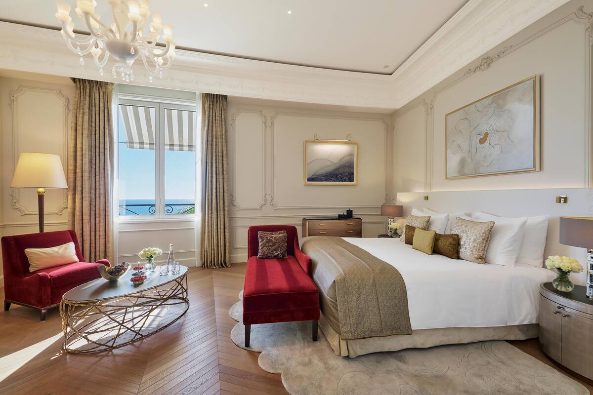 A hotel room with a bed, a red armchair, a small table with flowers, a desk, and a large window with curtains.