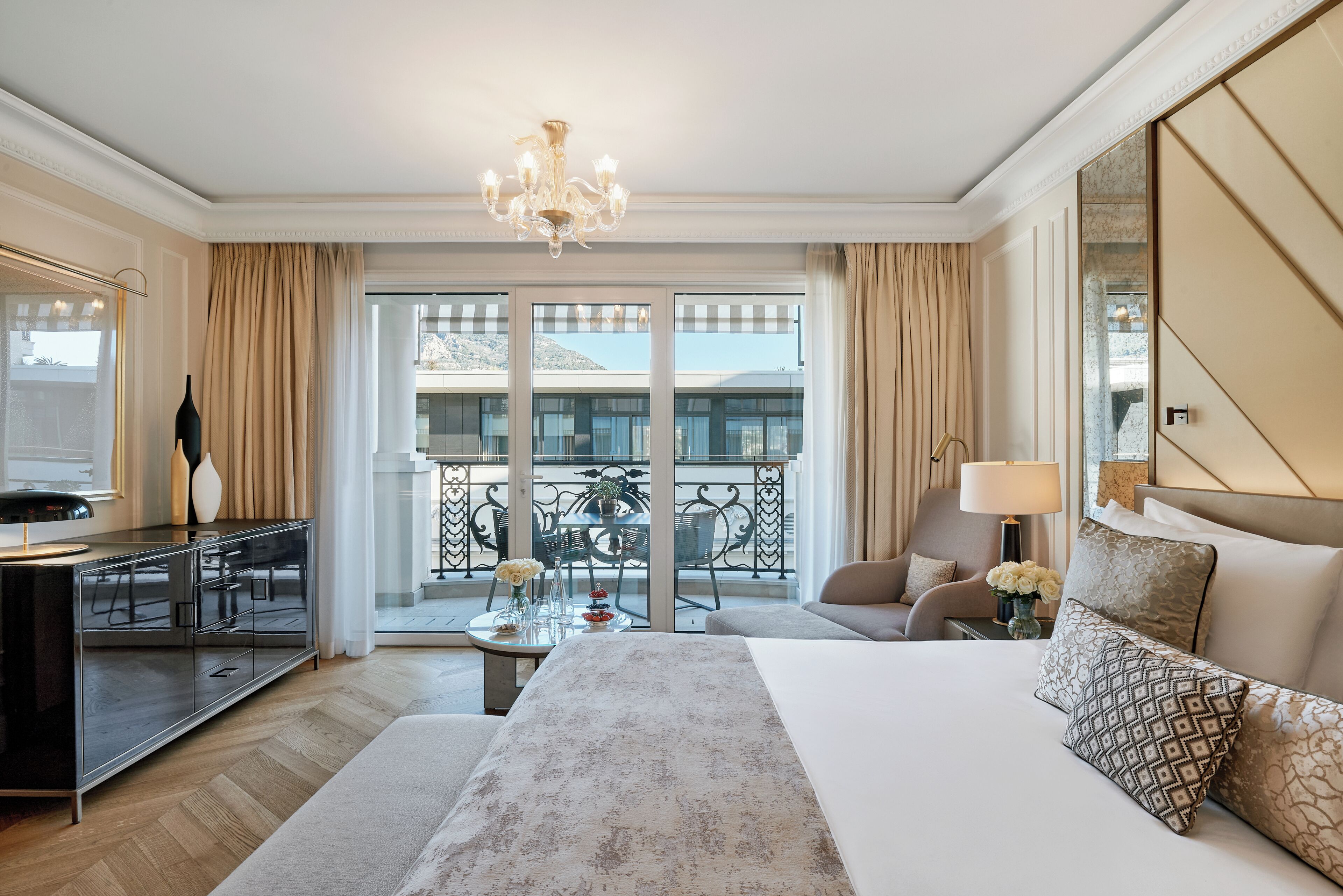 Habitación de hotel con una cama grande, un escritorio con lámpara, una silla, un balcón con mesa y sillas, y vistas al exterior.