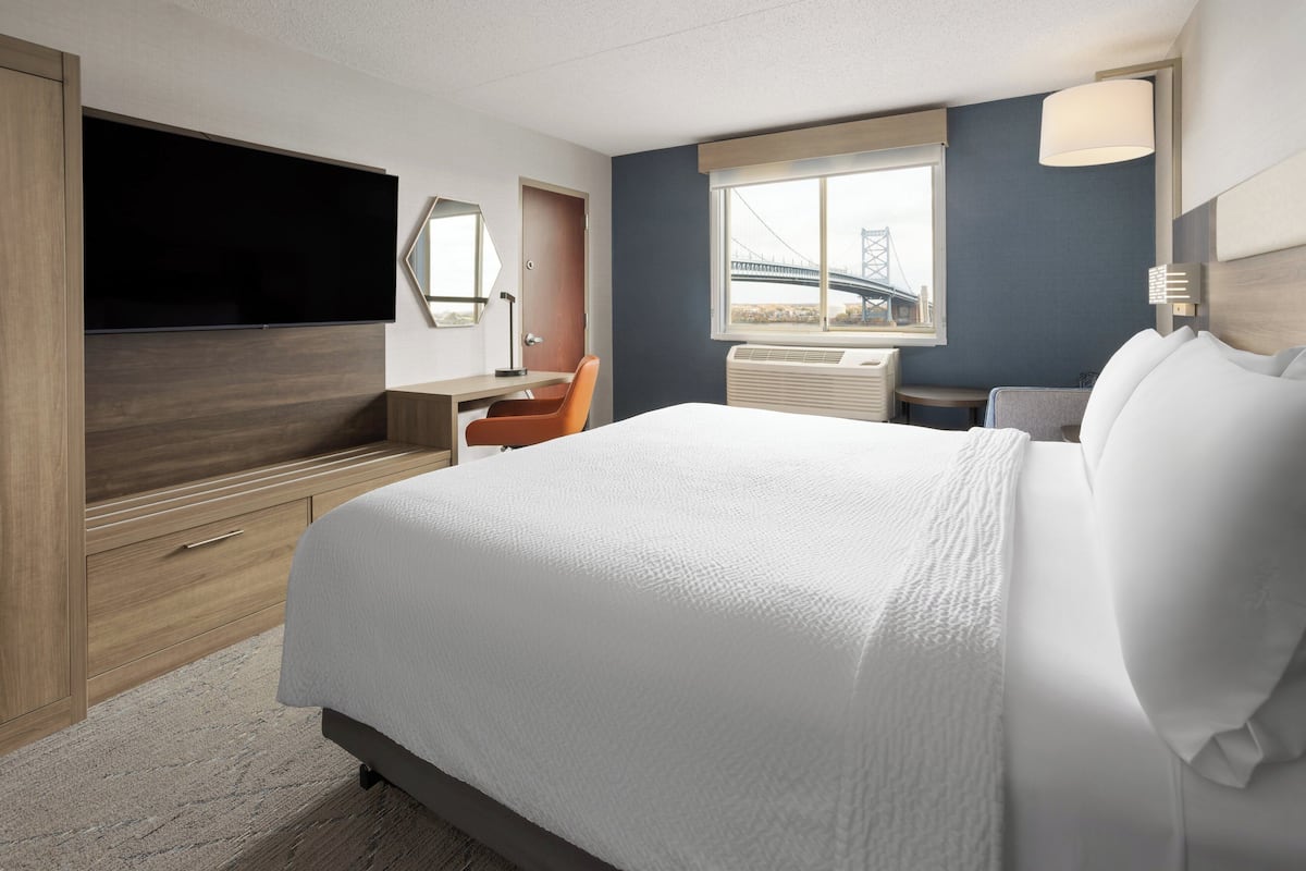 A hotel room with a large bed, a desk with a chair, a flat-screen TV, a window with a view of a bridge, and a wall-mounted lamp.