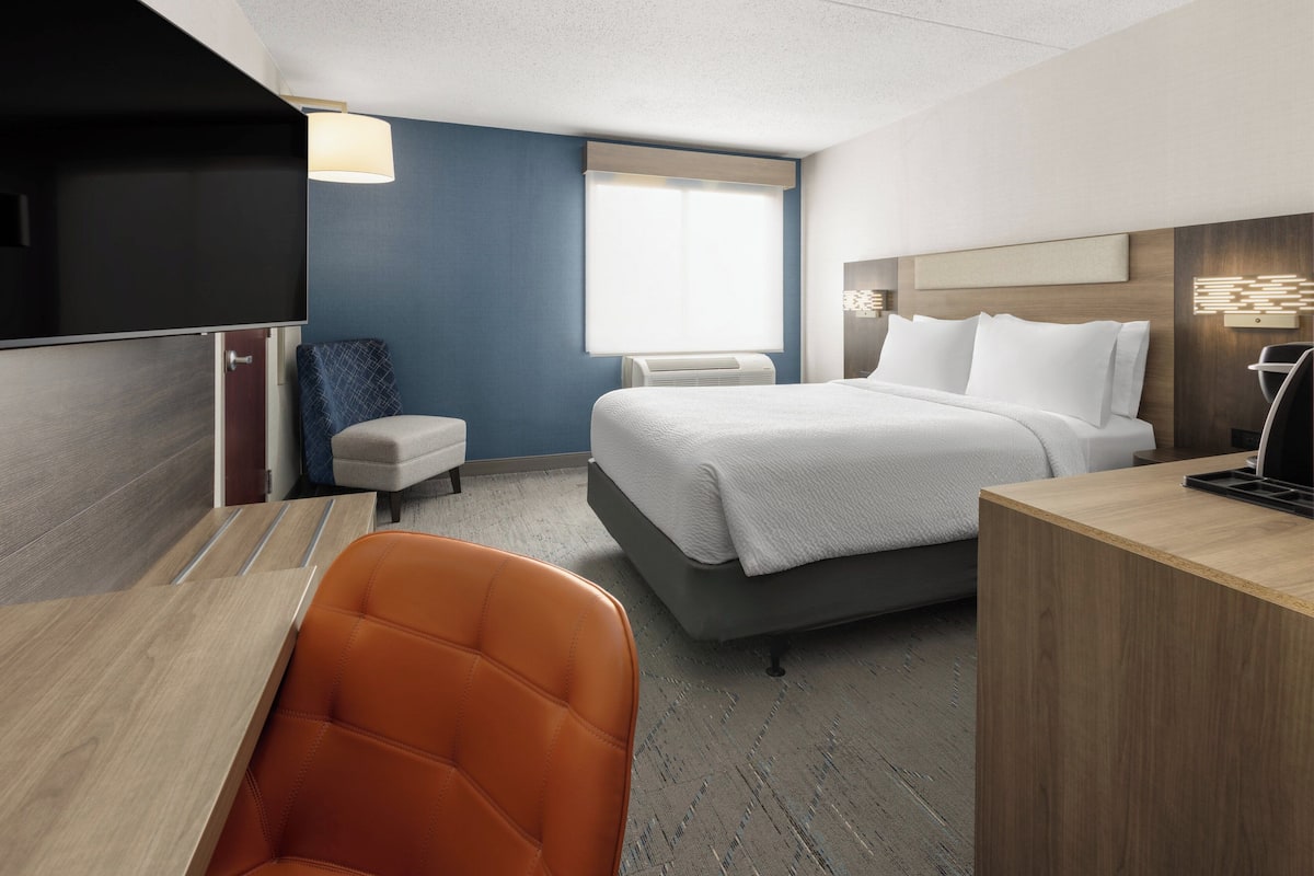 A hotel room with a bed, a desk, a chair, a small table, a lamp, and a window.