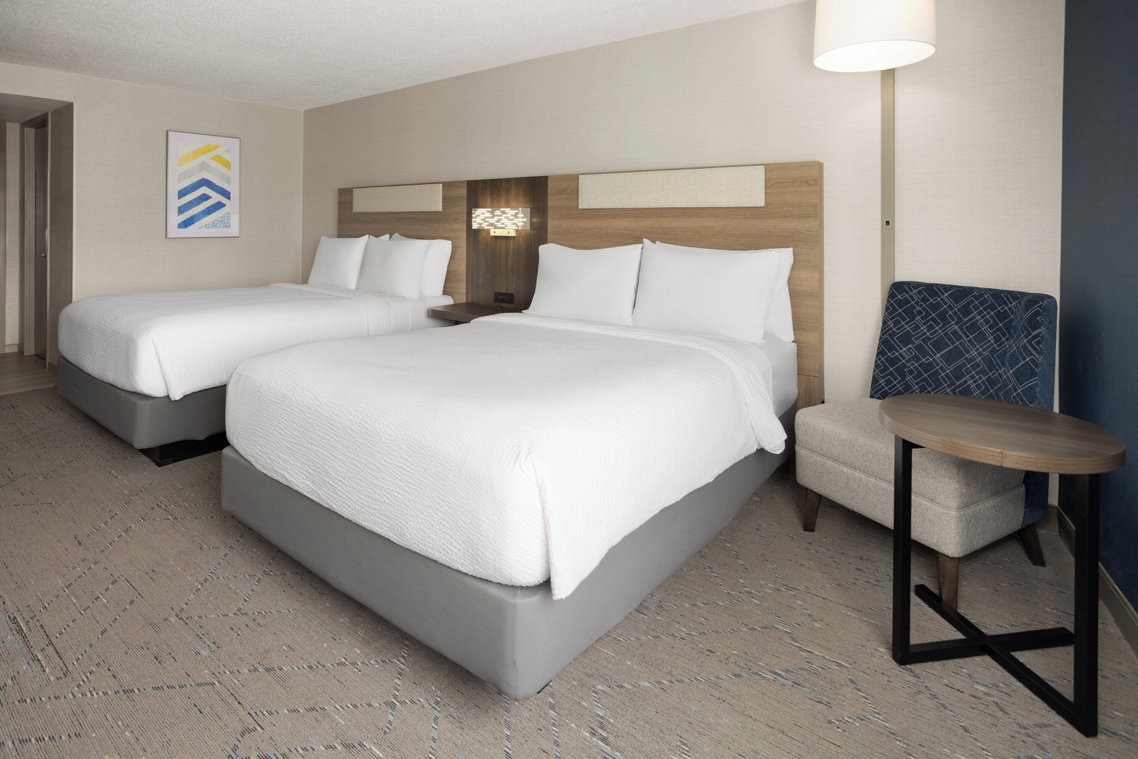 A hotel room with two beds, a nightstand, a lamp, and a small table.