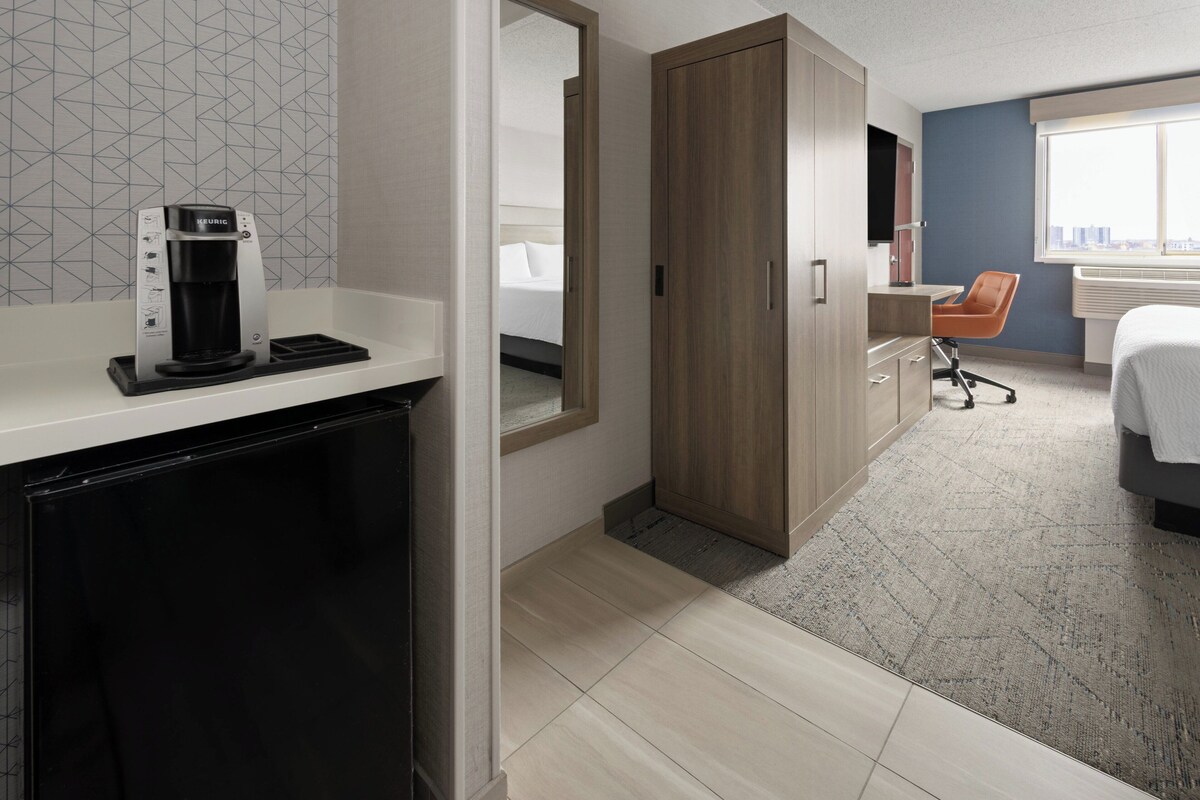 Quarto de hotel com cafeteira, guarda-roupa, secretária e uma cadeira laranja, além de uma cama.