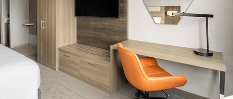 A modern hotel room with a bed, a desk, an orange chair, a TV, and a mirror.